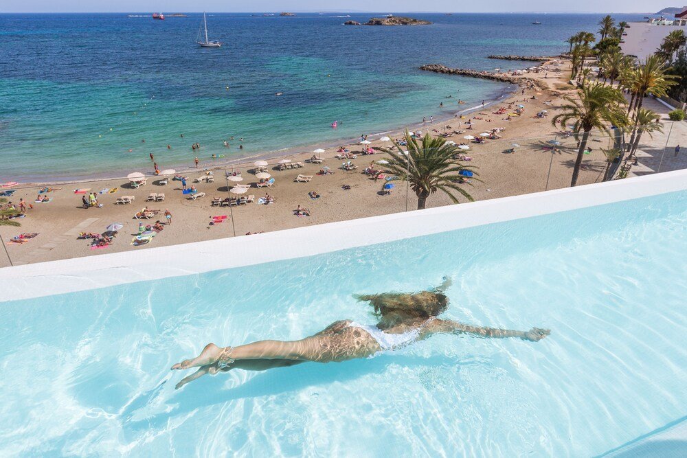 Pool with a swimmer and a beach with umbrellas beyond at One Ibiza Suites