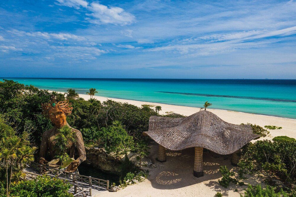 Beach view with a statue, a thatched umbrella and lush greenery at Palmaïa, Playacar